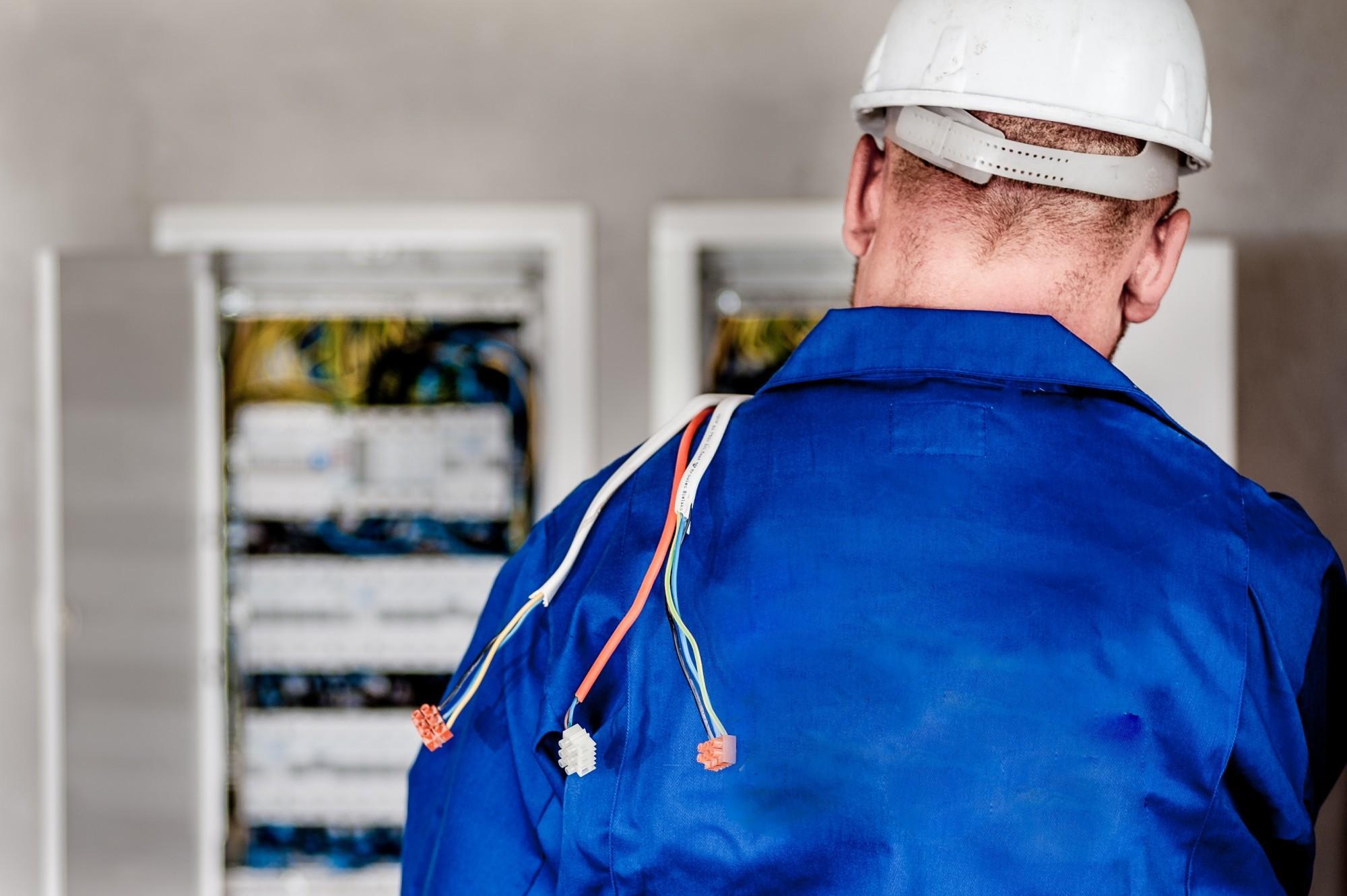 An electrician in a blue uniform and white hard hat stands by electrical panels with exposed wires draped over his shoulder, confidently demonstrating his expertise in handling complex wiring systems.
