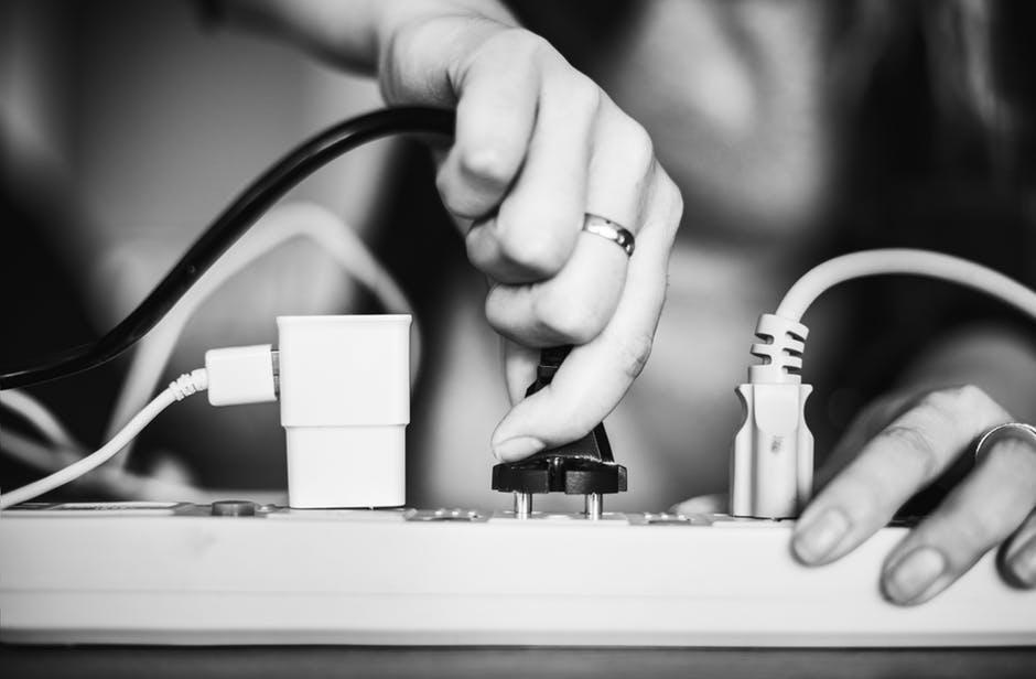 A person plugs a black electrical cord into a power strip that already holds other cables and a white adapter, just as electricians handle multiple connections at work.