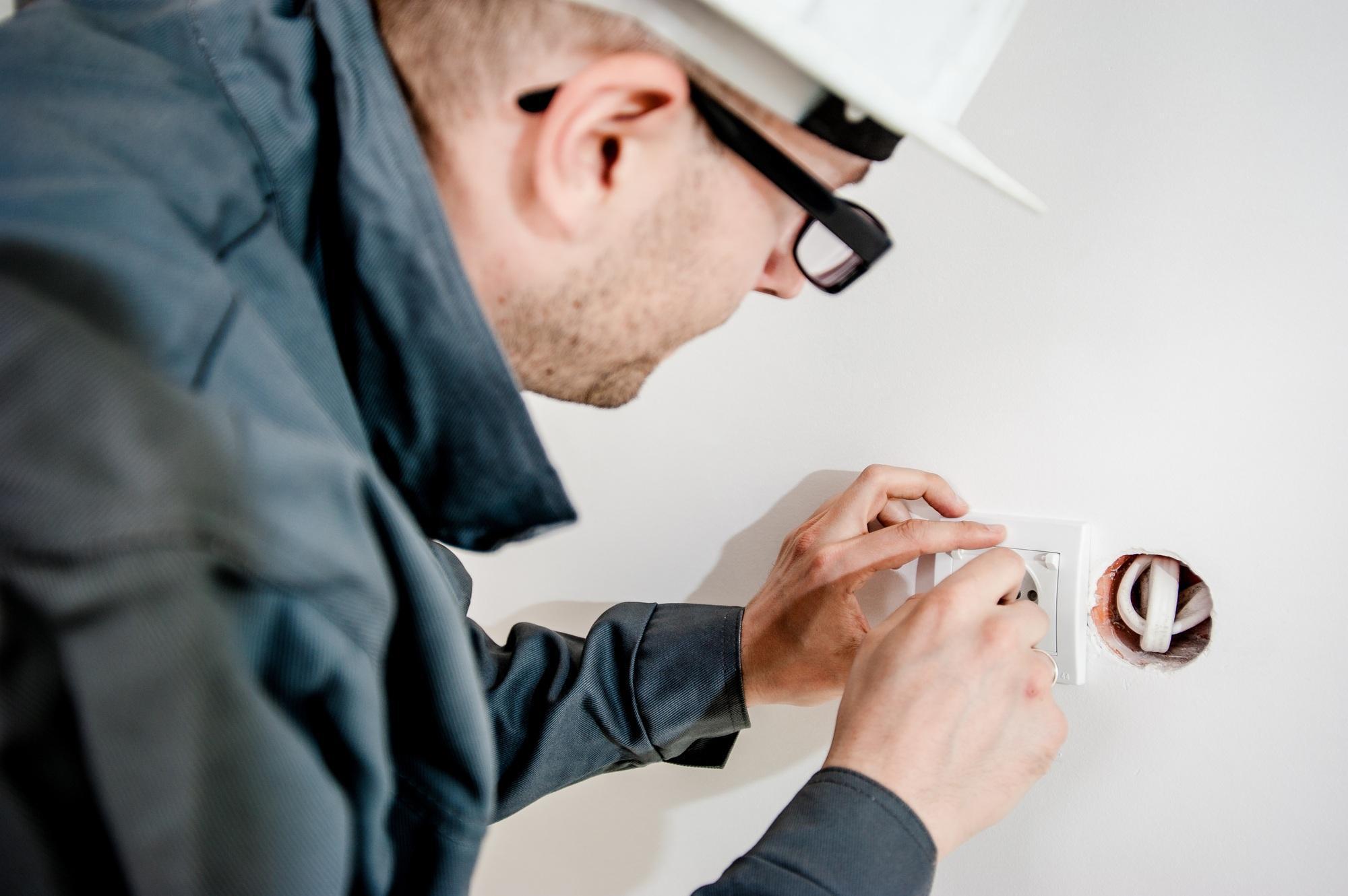 An electrician in a hard hat and glasses uses a screwdriver to install or repair a wall outlet.