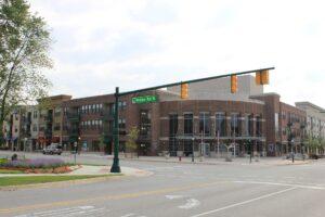 A modern, multi-story brick building stands at an intersection, with shops below, apartments above, and a Ridge Rd N street sign.