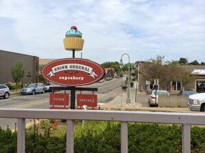 A Union General Cupcakery sign with a large cupcake illustration stands on a street, surrounded by buildings, cars, and greenery.