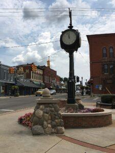 A street clock rises from a stone and brick base at a downtown intersection, surrounded by shops, traffic lights, and flowerbeds.