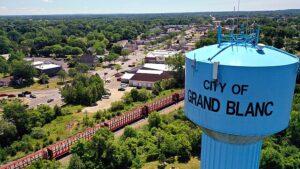 A blue water tower labeled "City of Grand Blanc" rises in the foreground, while a train, trees, and a small city fill the background.