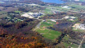 An aerial view shows a school campus featuring sports fields, a running track, wooded areas, roads, and a nearby lake.