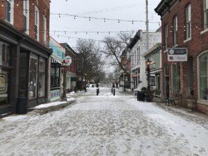 Snow dust covers the street as brick buildings, shops, and string lights fill a small town beneath a cloudy sky.