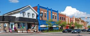 Shops and restaurants line a downtown street as cars park along the road under the sun. Colorful buildings show varied styles.