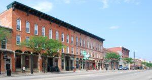 Shops fill historic brick buildings, while trees line the sidewalk and a clear blue sky stretches overhead in this street view.