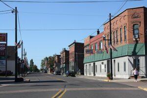 Brick buildings, parked cars, streetlights, and American flags line a small town main street as a person walks on the sidewalk.