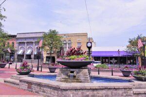 A sunny day lights up the brick plaza, where a central fountain and flower planters sit among small shops, flags, and a clock post.