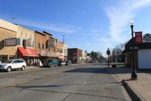 Cars line a small-town main street, parked by storefronts like a restaurant and pharmacy, beneath a clear blue sky.
