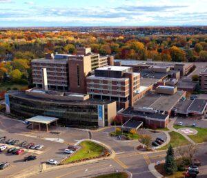 An aerial view shows a large hospital complex encircled by autumn-colored trees, parking lots, and roads.