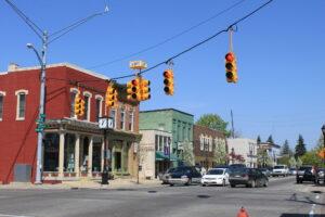 Traffic lights control a small town intersection as historic brick buildings and parked cars line the sunny, tree-shaded street.