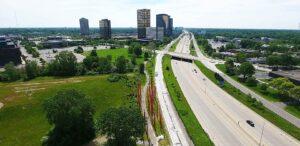 Aerial view shows a busy highway bordered by green space, a walking path, trees, and tall office buildings under a clear sky.