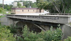 A concrete arch bridge with a decorative railing crosses Swartz Creek. A tan brick building and trees rise in the background.