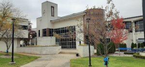 A modern building displays a clock tower and large windows. Trees, shrubs, and blue ribbon decorations fill the landscaped grounds.