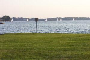 A grassy lawn stretches to a calm lake where several sailboats float. A small sign stands by the water and trees line the far shore.