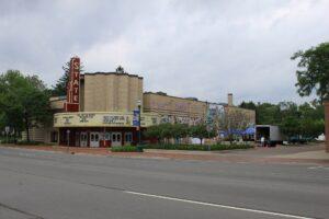 A vintage-style theater with a "STATE" marquee sits on a corner, featuring a mural, nearby trees, street signs, and a cloudy sky.