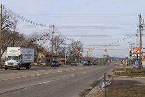 Several vehicles travel down a wide street lined with leafless trees, passing traffic lights, utility poles, and roadside businesses.
