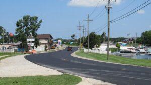 A newly paved road winds through Whitmore Lake, passing power lines, parked cars, boats, and buildings under a clear, sunny sky.