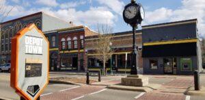 You see Depot Town’s street with a post clock, historic brick buildings, a large sign in front, and businesses along the sidewalk.