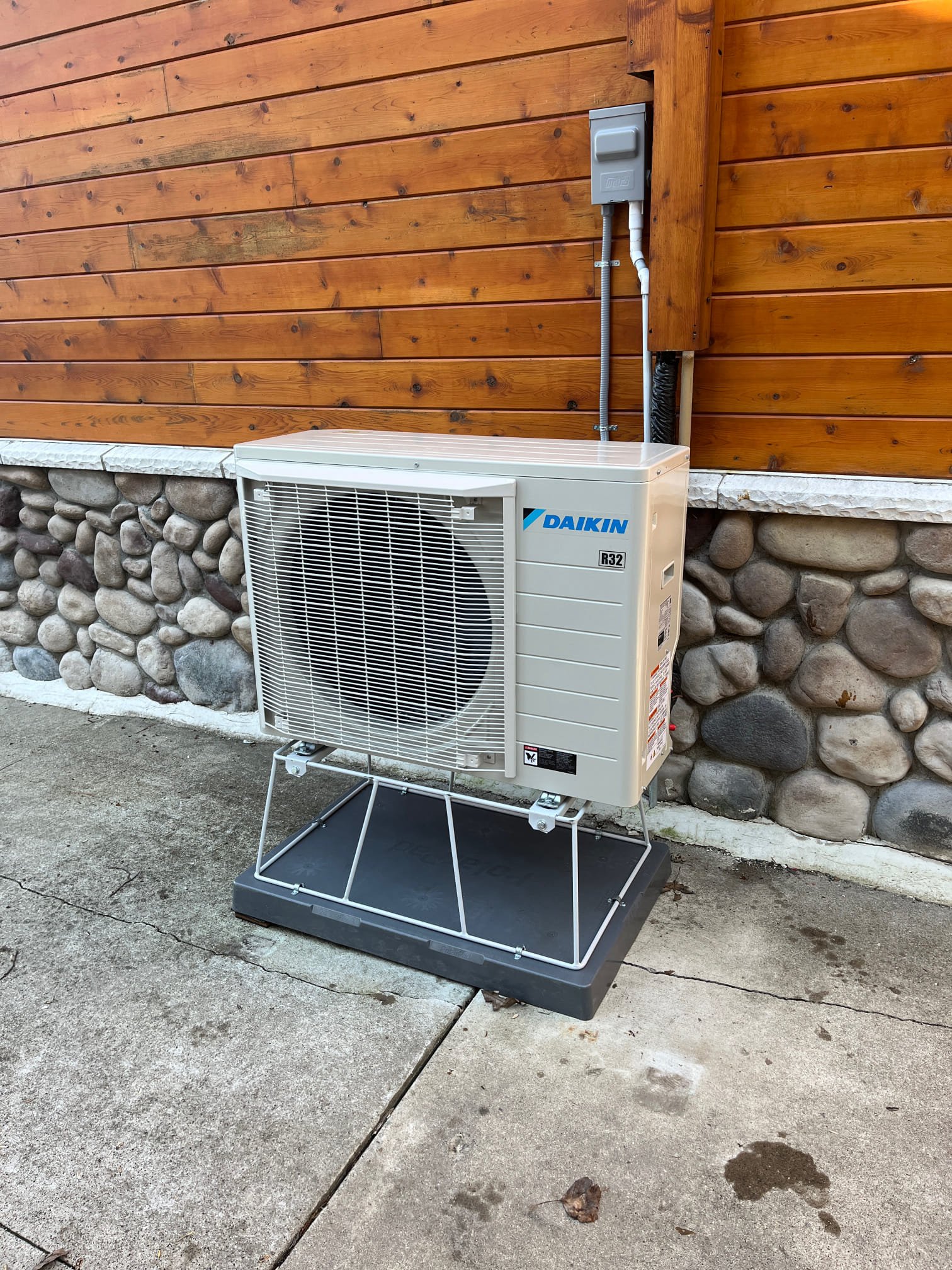 A Daikin outdoor AC unit sits on a metal stand beside a stone and wood building, showing how to install one in Michigan.